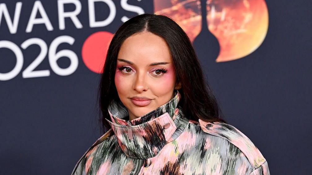 Jade has long black hair, she is wearing a high neck green and pink pattern jacket.