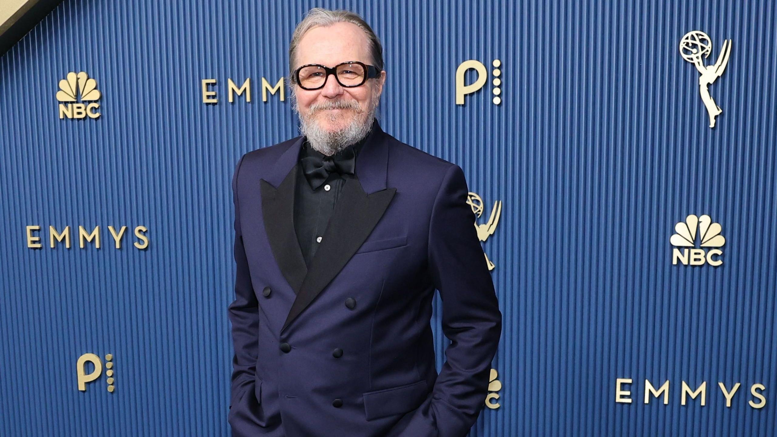 Gary Oldman has short grey and brown hair and grey facial hair. He is wearing a black shirt with a black bow tie and a navy blue suit jacket. He is standing in front of a blue background with gold signs that read "NBC" and "Emmys".