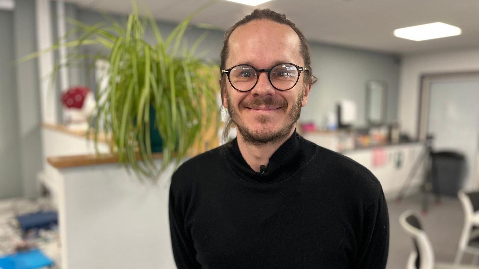 Darren is wearing a black jumper and dark-rimmed glasses. His hair is tied back from his face and he is smiling at the camera. Behind him are desks and office furniture and a large plant on a shelf.