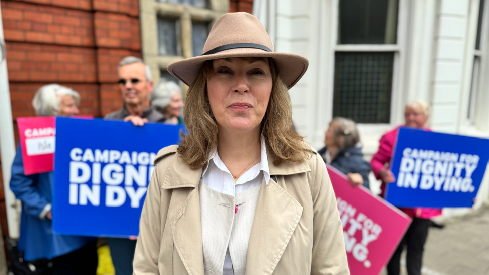 Sarah Wootten is wearing a white shirt, cream coat, and beige hat. She has shoulder length blonde hair and is standing in front of campaigners outside Tynwald with Dignity in Dying placards.