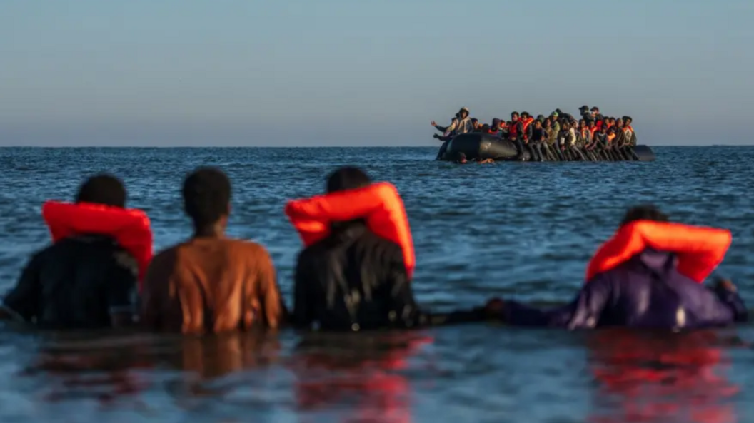 A group of migrants are seen in the distance on an inflatable boat in the Channel. In the foreground are four people in the sea wearing life jackets.