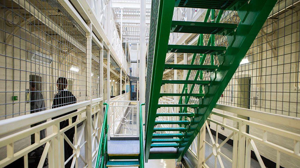 A prisoner is escorted to his cell by an officer next to a set of green stairs situated in between rows of cells on Benbow wing inside HMP/YOI Portland.