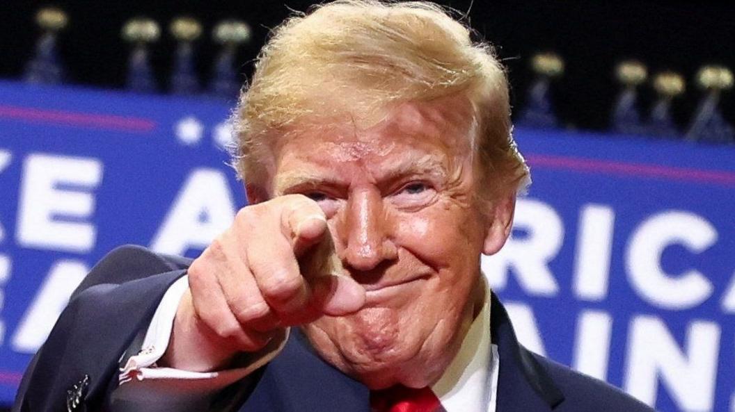 US President Donald Trump smailes and points at the camera against a blue and white backdrop at a rally