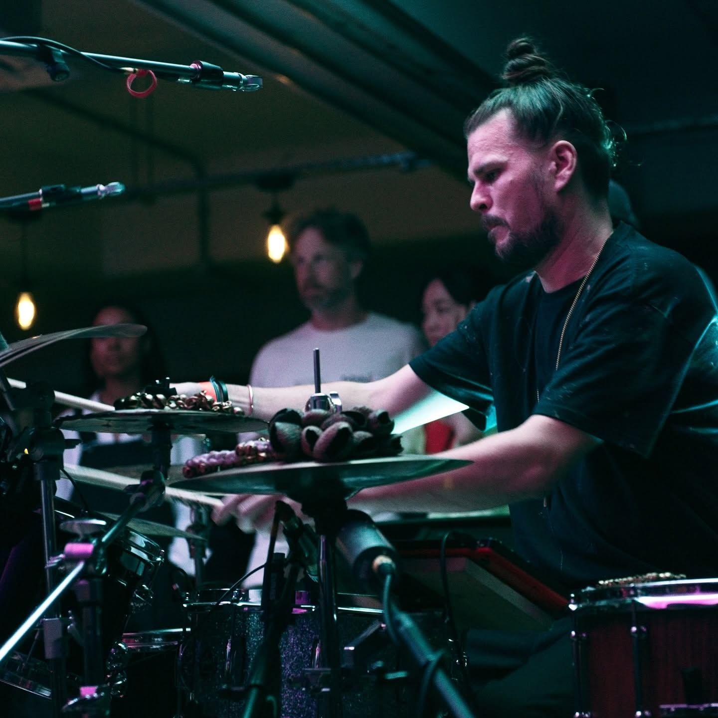 A man with a hair bun sitting at a drum set. He is wearing a black t-shirt and on a stage with purple and blue lighting.