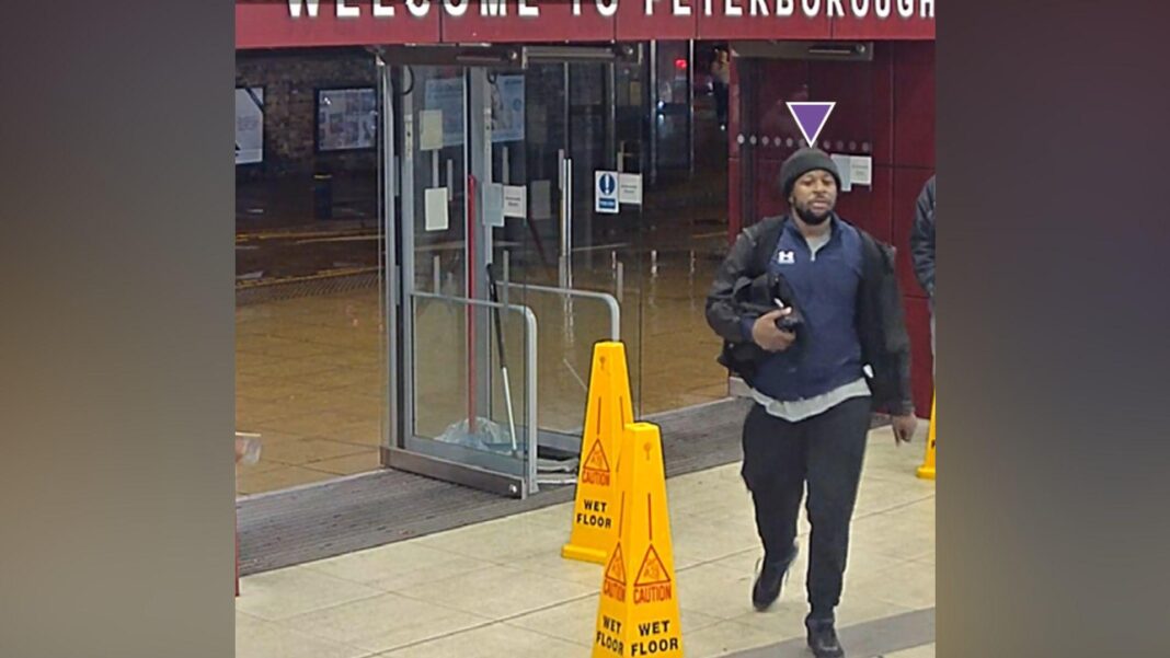 A man in dark clothing and black woolly hat carries a small black bag under his right arm. He is outside glass doors at a railway station and is walking past yellow cones that say 