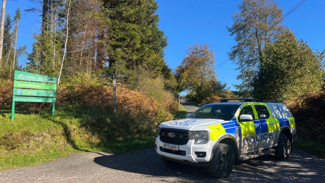 Man, 80, who died in light aircraft crash near Loch Lomond named Police car blocks off road with tall trees on either side.