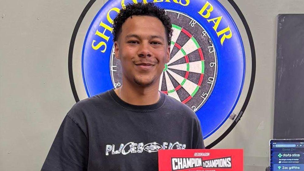 Zac Griffiths stands in front of a dartboard that reads "Shooters bar". He has dark brown hair and is smiling. He holds a red sign reading "Champion of Champions".