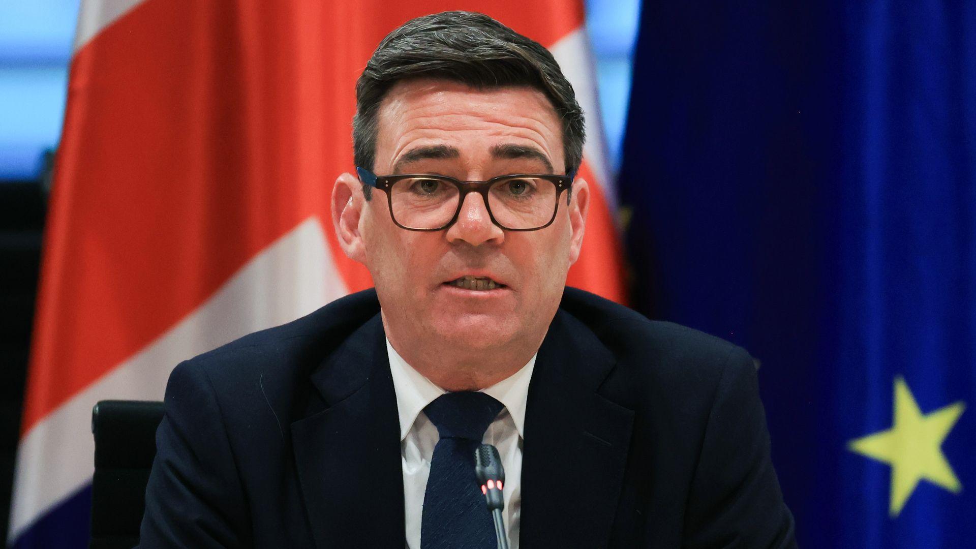 Andy Burnham, UK prime minister, sits in front of a microphone with a British flag and an EU flag behind him