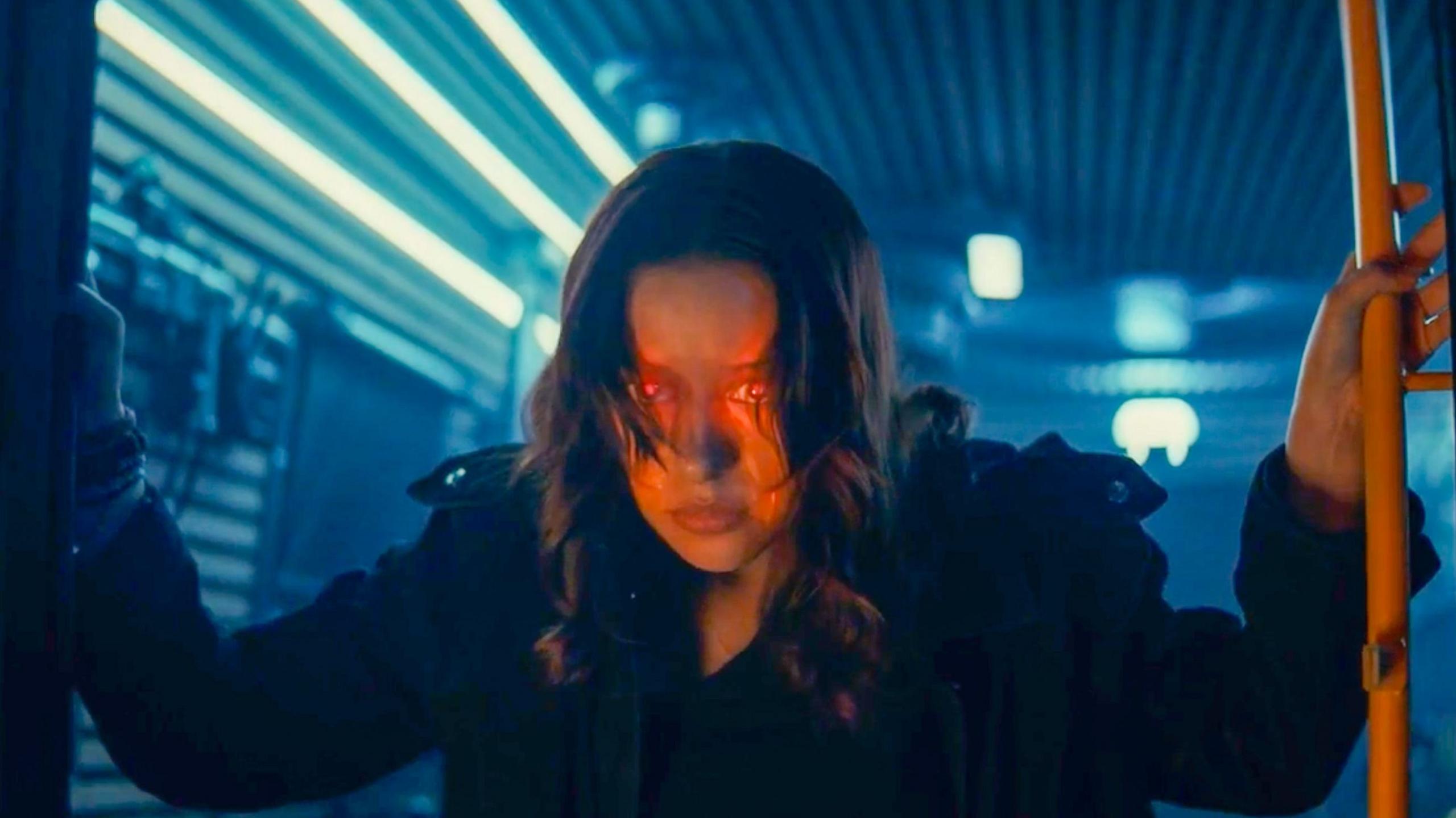 A woman with a dark coat on and dark brown hair. She has red glowing eyes and she looks out.