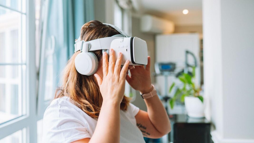 Dread presenting at work? Read these tips before your next meeting A woman with brown hair using a white virtual reality headset piece, she is wearing a white top and has her hands on the set. The background has a big window on the left and a black table with a potted plant on top on the right.