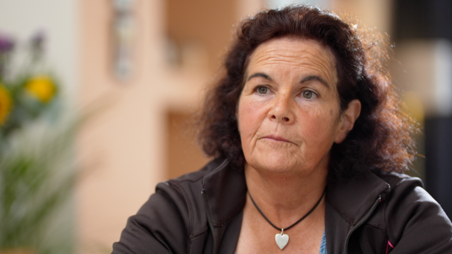 Linda Williams looks beyond the camera with a serious expression. She wears a silver heart pendant and dark coloured top and her hair is dark and curly.