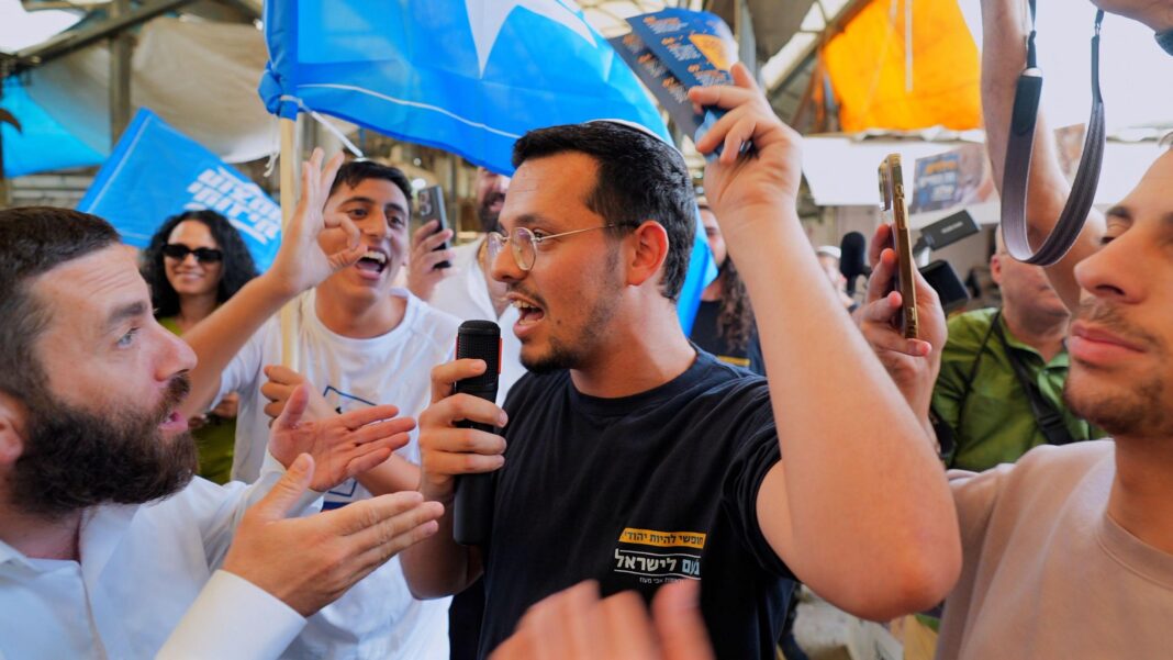 A man in a navy t-shirt talks into a microphone surrounded by several other men waving flags, in Ramle market, Israel