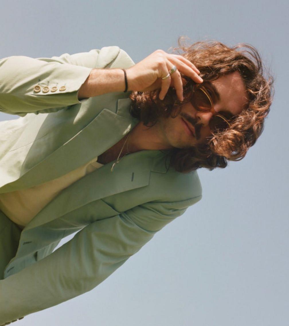 J Casa - a man with long brown hair and a moustache touches his sunglasses while bending down towards the camera. He is wearing a light green uit and a light yellow v neck top.