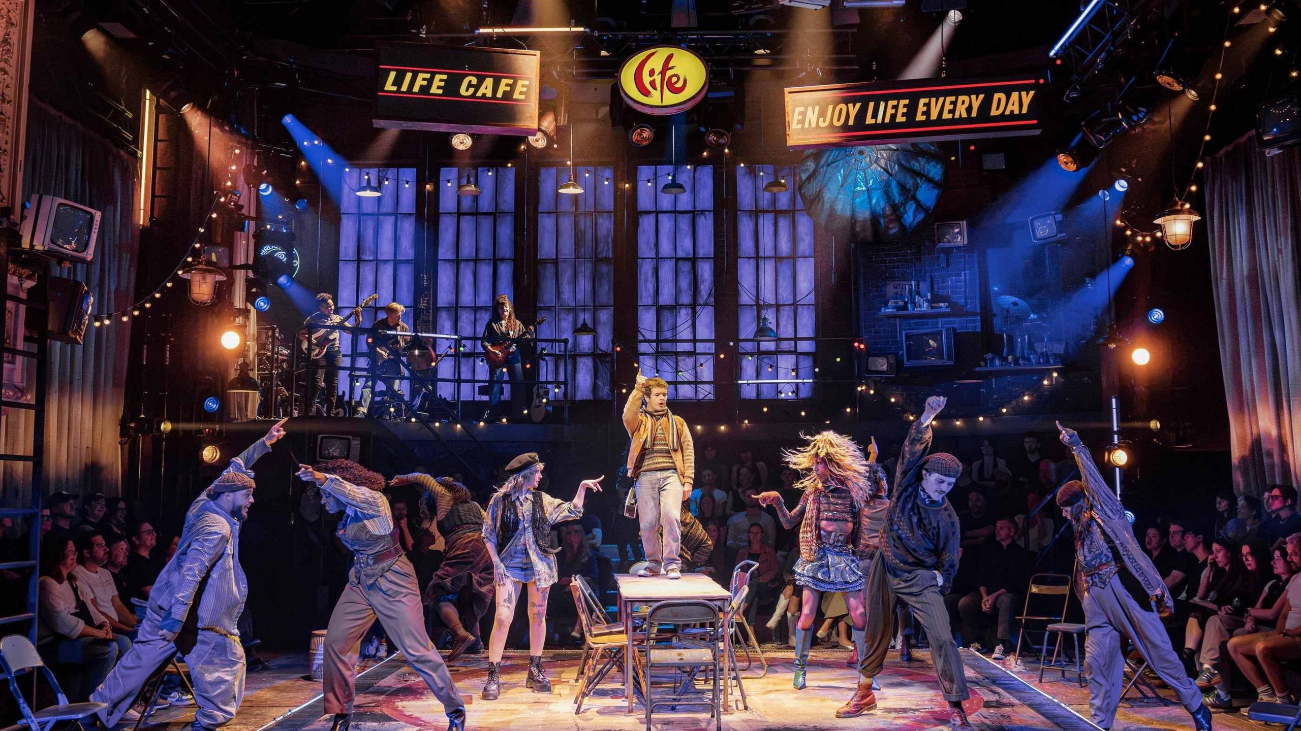 The cast of Rent mid-performance on stage. Gaten Matarazzo stands in the middle of the table singing, while the ensemble dances around him. There are fairy lights and a band in the background, while the audience watches on