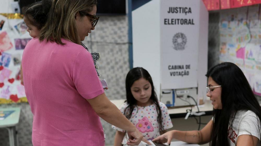 A voter submits a ballot at a polling station in Brazil as an election worker assists. A voting booth marked “Justiça Eleitoral” stands nearby