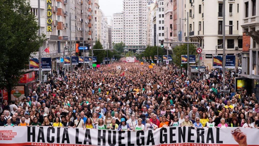 A large crowd taking up the entirety of a wide road in Madrid. The people at the front hold a large sign which reads 'hacia la huelga general'