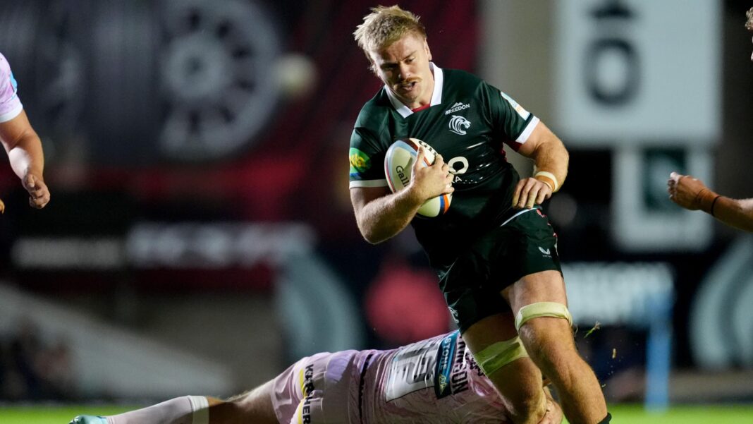 Aaron Wainwright in green and white Leicester kit and with bandages above both knees runs with ball in hand as he avoids a tackle by a player on the ground in pink.