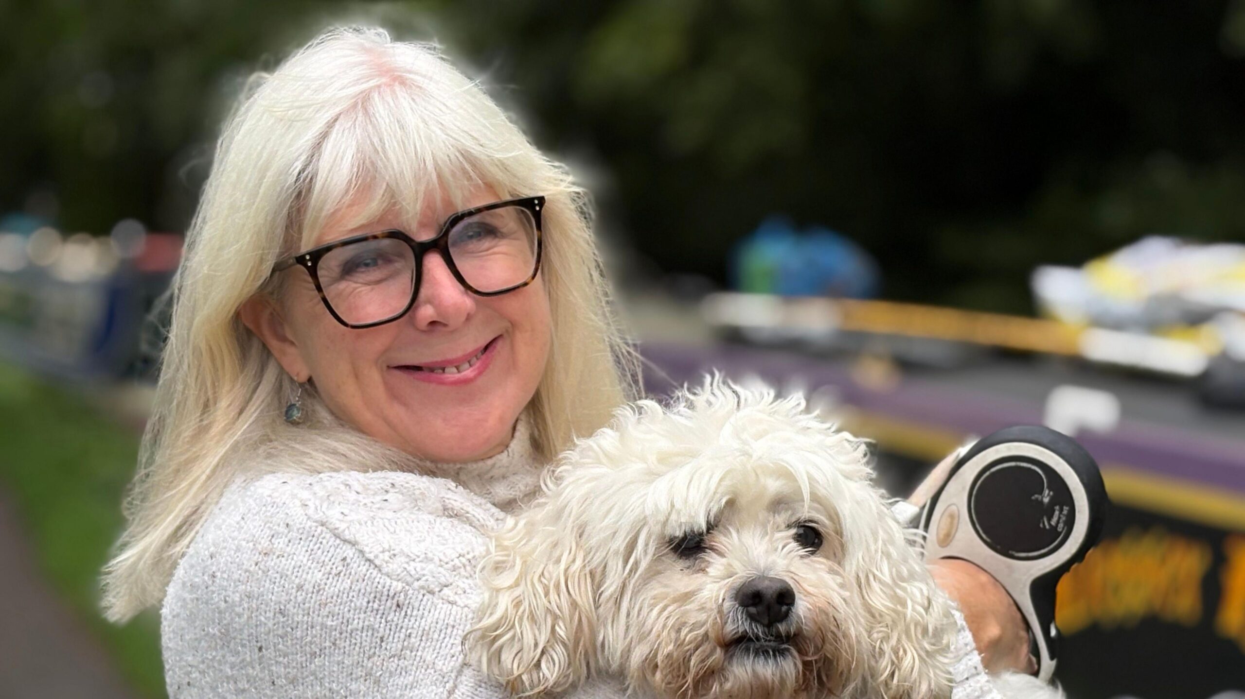 A woman with shoulder length blonde hair and glasses smiling at the camera. She is holding a small white dog in her arms that has a curly coat.
