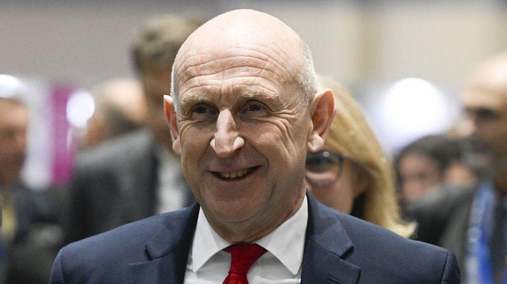 Head and shoulders of John Healey in a crowd, wearing suit and red tie