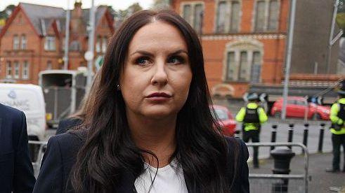 A woman with long brown hair. She is standing outside, and is wearing a white top and a dark blazer. In the background, there are a number of police officers.