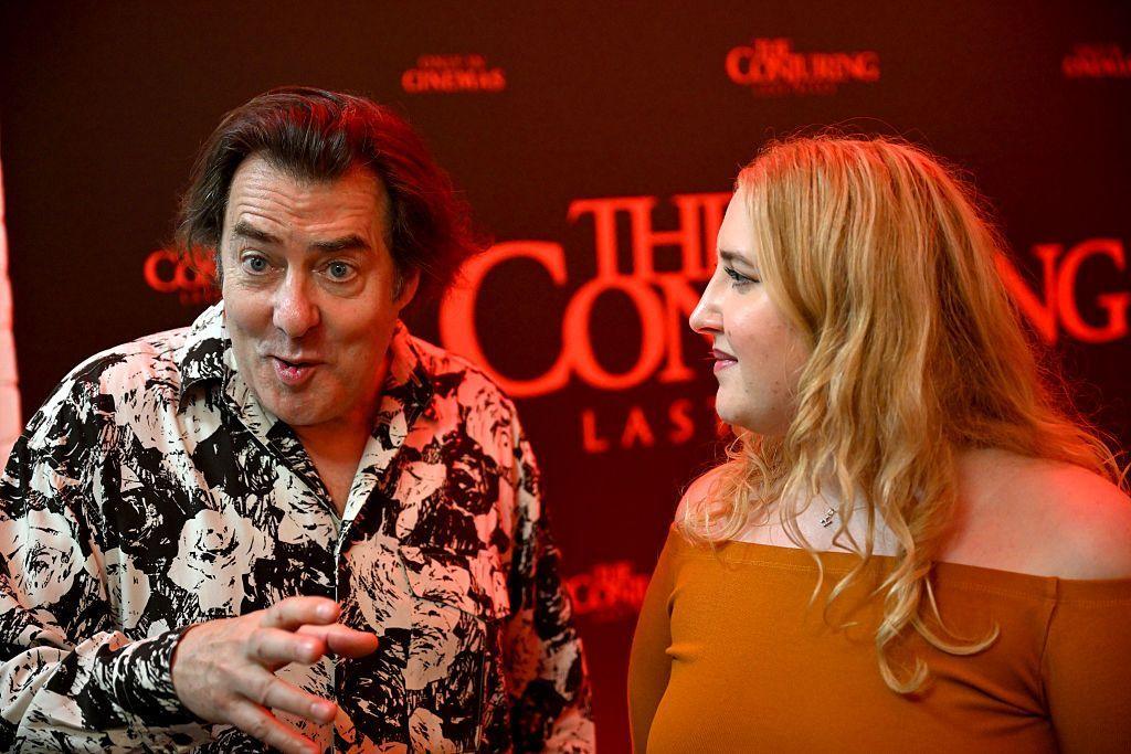 Tv presenter Jonathan Ross, with a wide-eyed expression looking at the camera with hand out in front of him. He is wearing a black shirt with a white floral pattern. His daughter Honey, wearing an orange dress cut-off at the shoulders is looking at him with a smile on her face