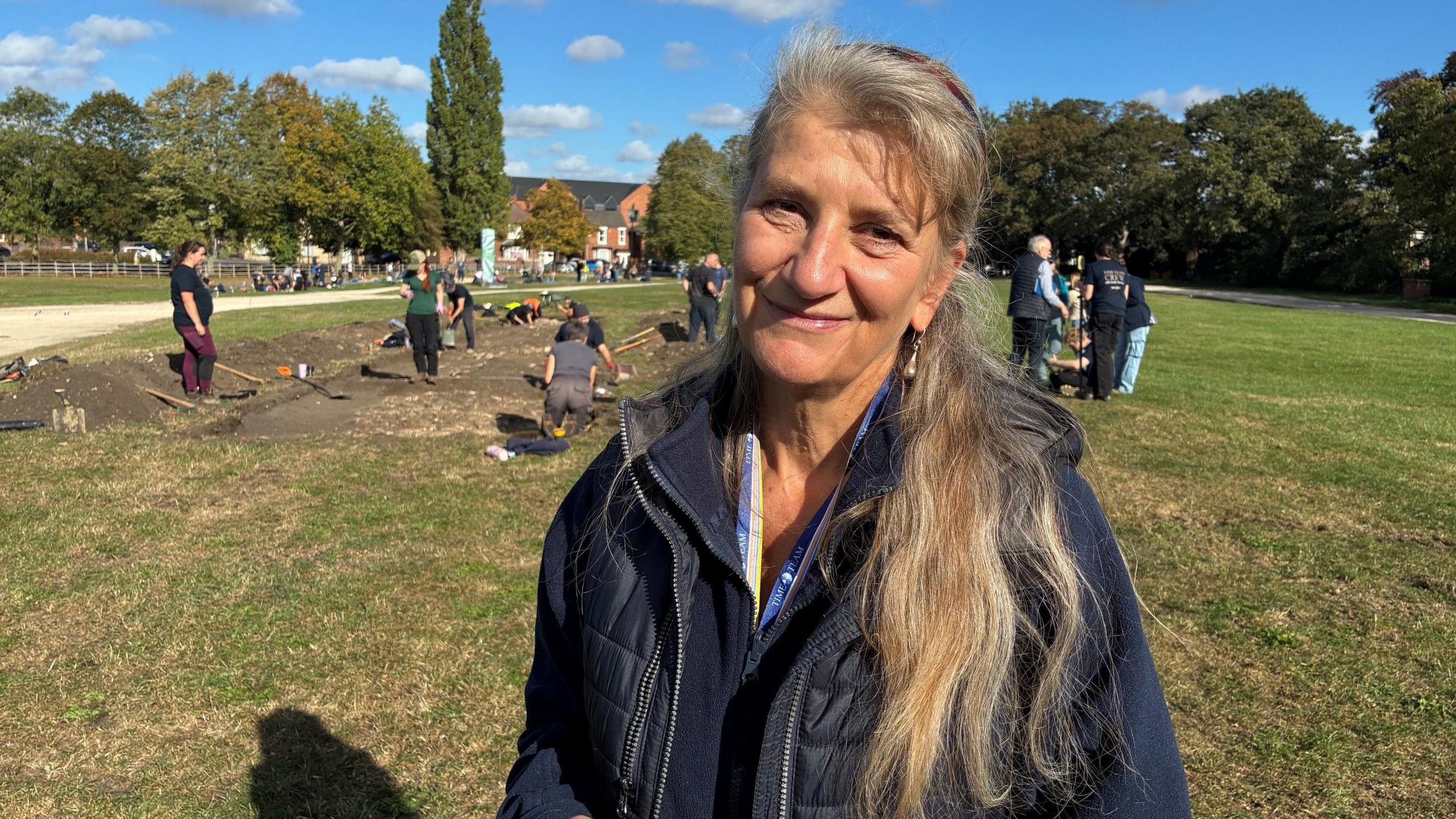 Carenza wears a navy jacket and smiles for the camera in front of the pit where people are digging behind her. She has long blonde hair which is held back in a ponytail.
