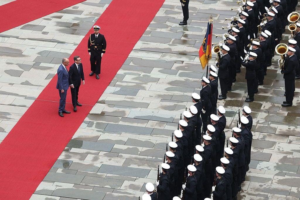 Burnham and Merz stand on a red carpet honoured by three rows of military personnel including a military band.