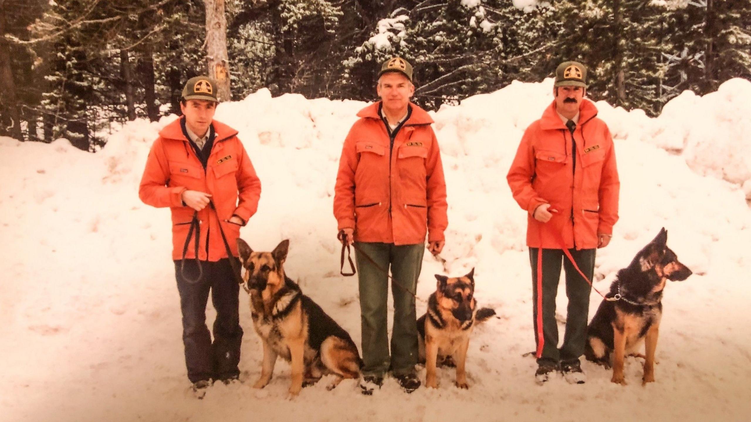 An image showing three dog handlers as part of the Canine rescue team, with their dogs beside them. All three dogs are German Shepherds, and all three handlers are dressed similarily in a bright orange jacket, a park warden hat and dark trousers.