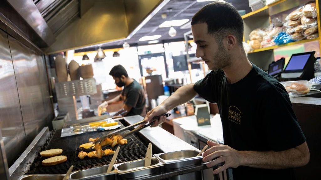 A young-looking worker uses tongs to flip meat on a grill.