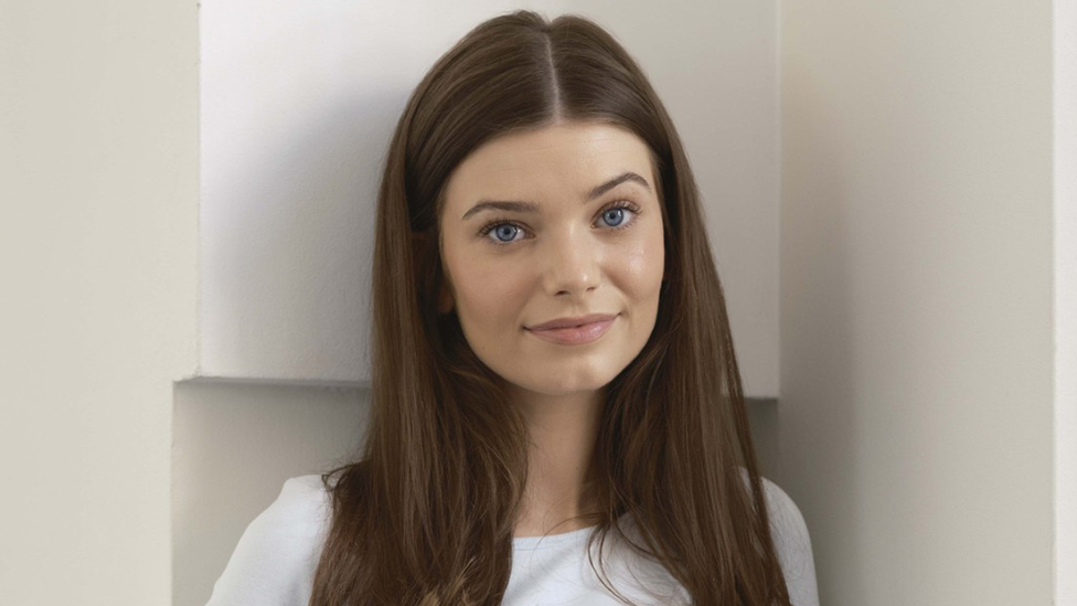 A close-up portrait of Leisa looking directly at the camera. She has straight brown hair reaching past her shoulders, which is parted in the middle. She stands indoors with a plain, light-coloured background.