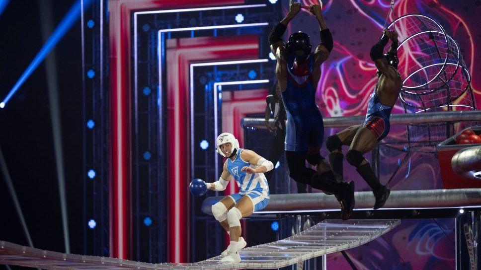 Josh Mcdonald, wearing a white-and-light-blue athletic uniform, helmet, and protective pads, balances on a narrow transparent walkway while holding a blue ball.   Two Gladiators  in dark blue and red outfits leap overhead from a higher platform trying to knock him over.