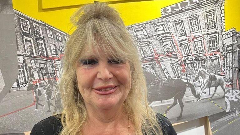 A smiling fair haired woman with long hair and a black top stands in front of black and white art which features horses on a street in front of brick buildings