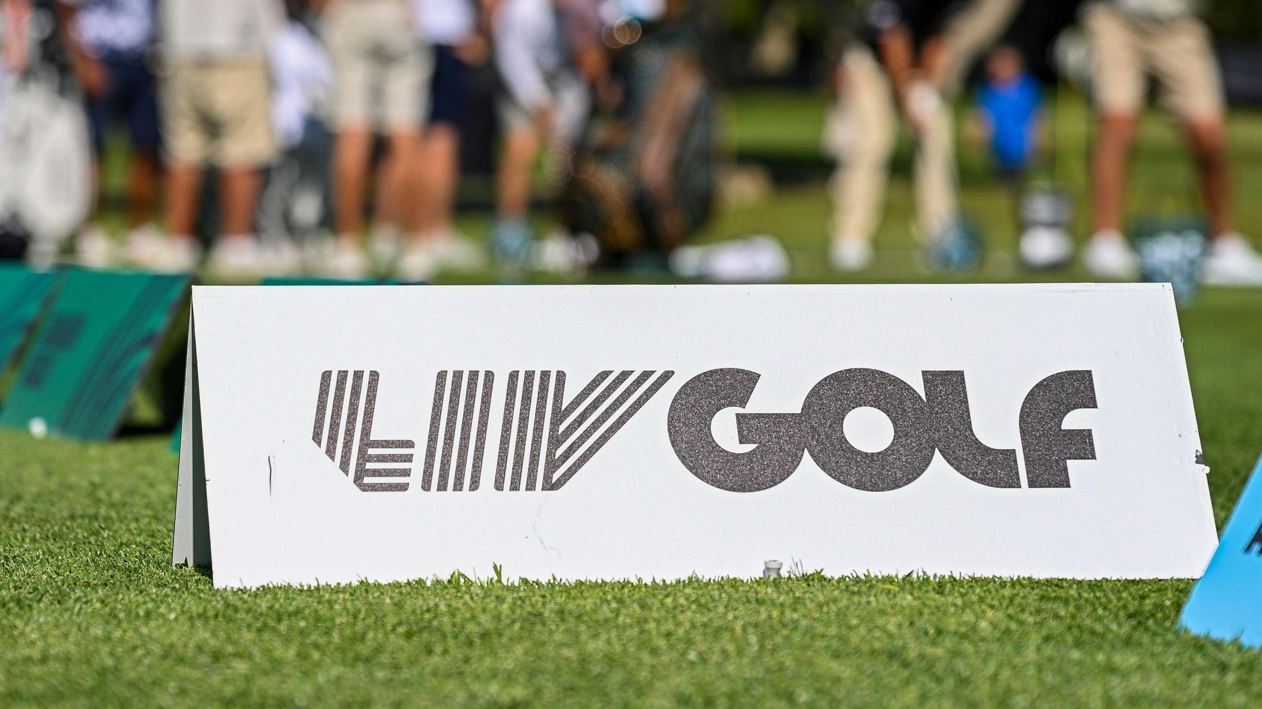 A marker with LIV Golf branding on some grass, with golfers out of focus in the background