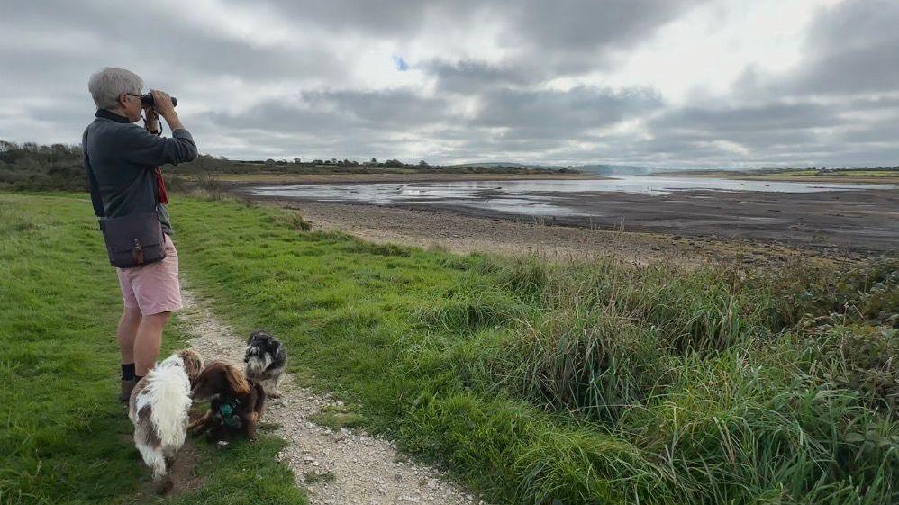 A man is standing on an area of grass overlooking a reservoir. Three dogs are standing by his feet. The man is is looking through binoculars. Water levels are low and mud is visible.