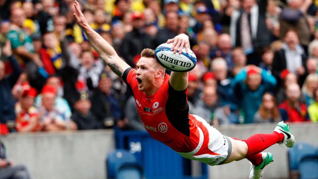 Chris Ashton performs a swan dive while scoring a try for Saracens. He is holding a rugby ball in his left hand and diving for the line, wearing a red shirt, white shorts and red socks