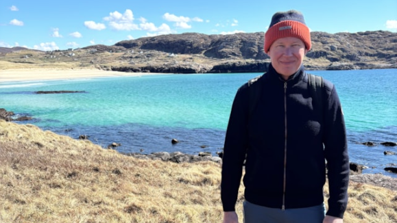 Michael Mullins standing in front of a blue lake in black overcoat and red beannie hat