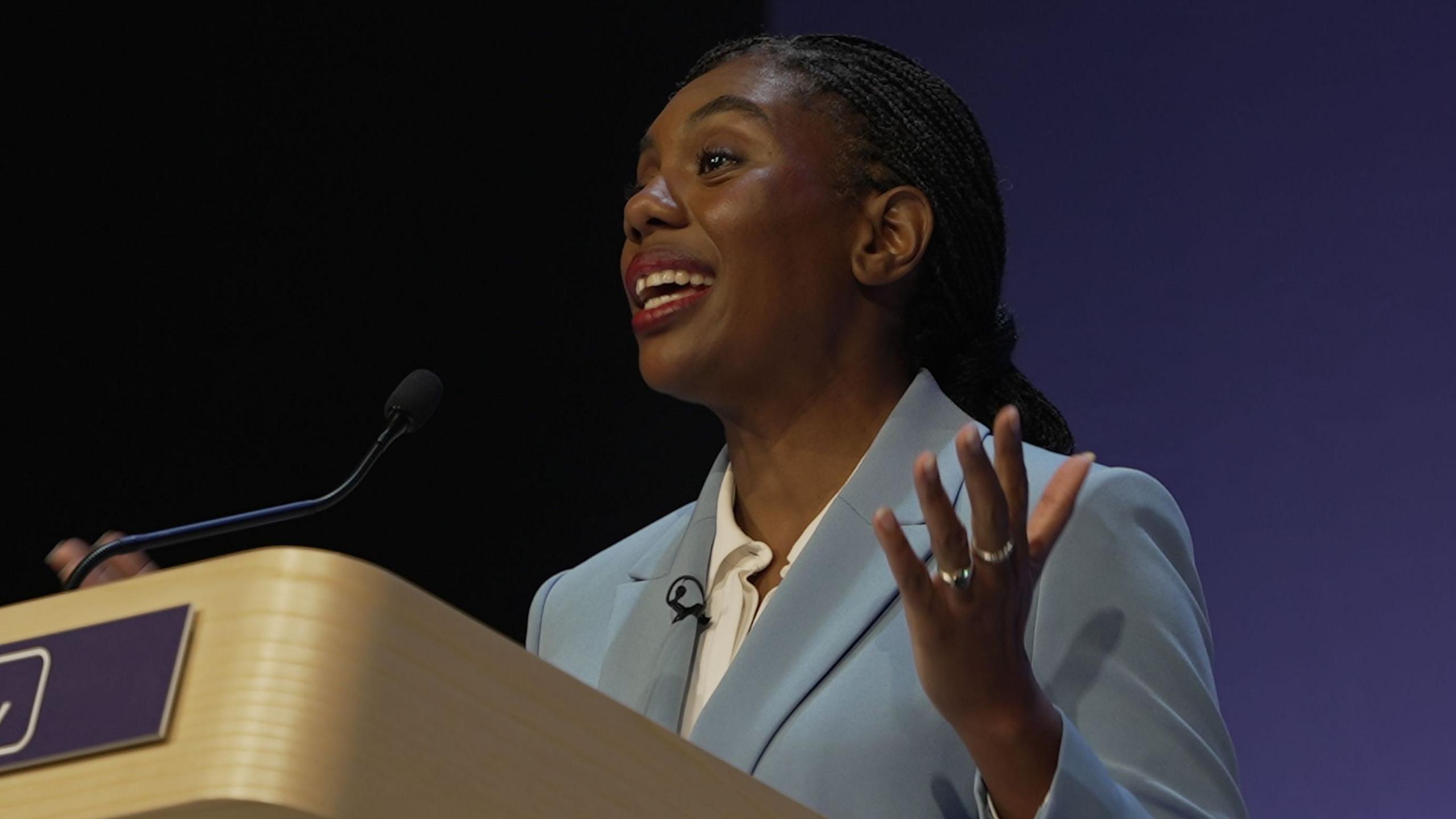 Kemi Badenoch stands at a podium gesturing with her hands, wearing a light blue suit jacket over a white shirt