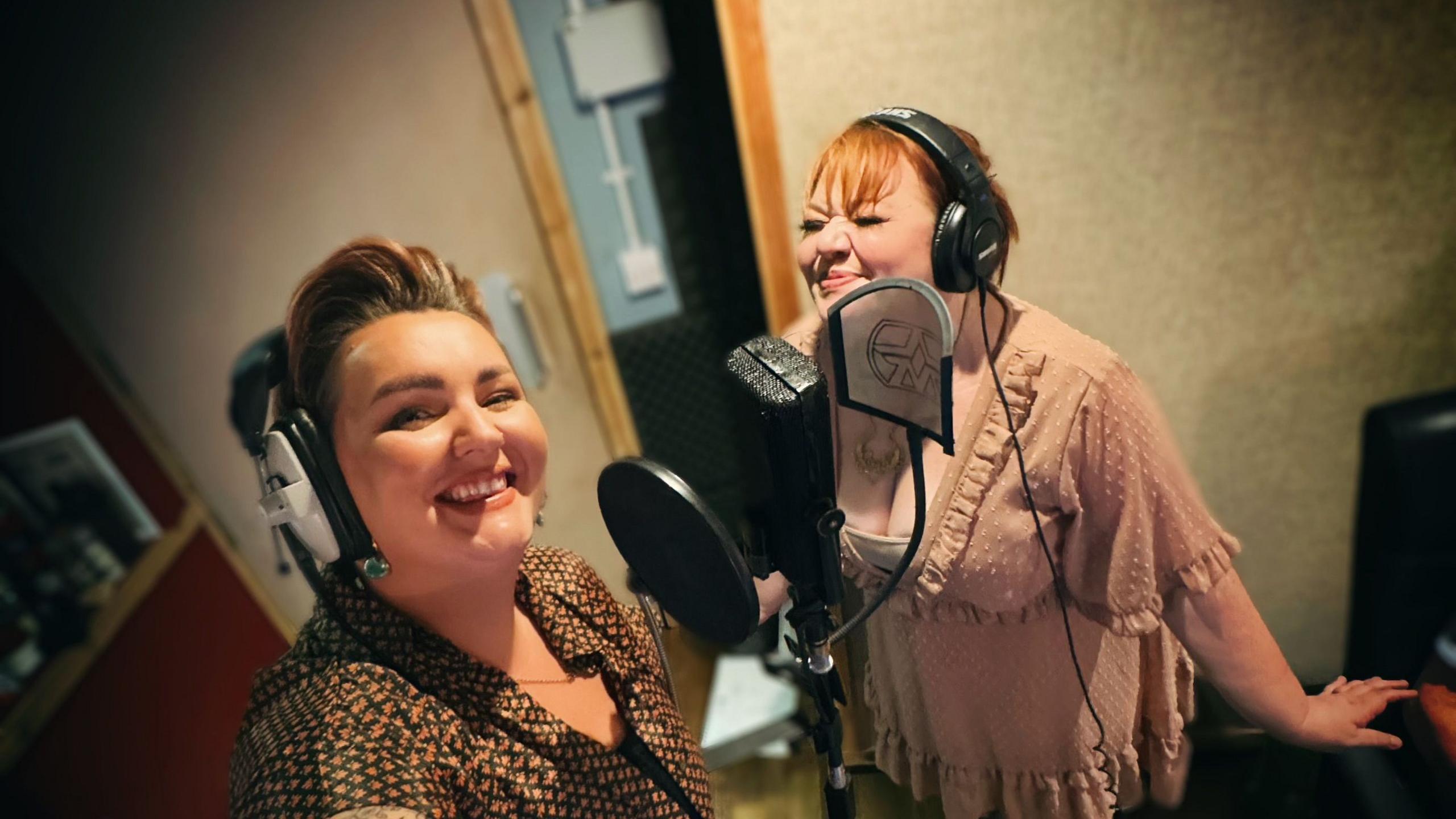 The two women singing in a studio. There is a mic in front of them and they have headphones on.