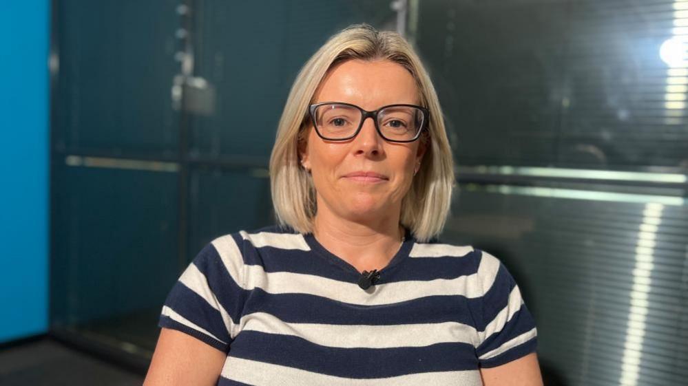 A woman with shoulder-length blonde hair wearing black glasses and a striped navy and white top looks directly at the camera. She's sitting in a room with metallic grey blinds behind her and a bright blue wall to the left.