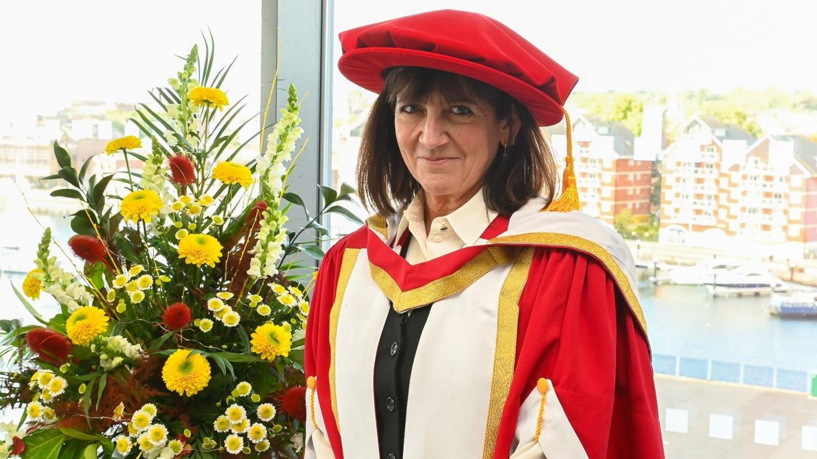 Emma Freud wears a red cap and a red, white and gold gown over a black jacket. She is smiling and stands in front of a window, next to a plinth with flowers. The view is of Ipswich Marina and the Waterside is filled with boats.