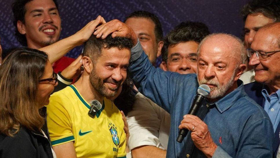 André Janones, wearing a yellow Brazilian football shirt, smiles as President Lula put his right hand on top of his head in front of other jubilant supporters. Lula is wearing a blue denim shirt and holds a microphone in his left hand, which has a missing little finger.