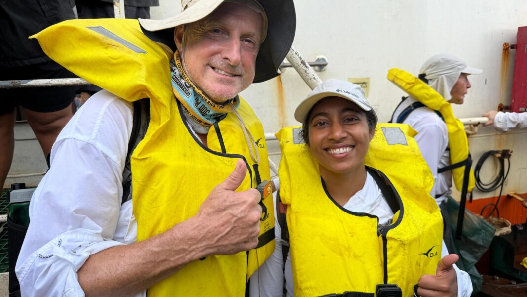 Search for Amelia Earhart’s plane resumes after stormy weather Expedition members Steve Schultz and Sirisha Bandla give a thumbs up and smile to the camera while wearing yellow life vests and white tops on the deck of a ship. Schultz wears a cream bucket hat and Bandla a cap.