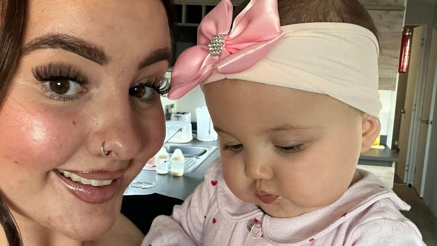 Sophie Price (left) is a young woman who has brown eyes and brown hair. She is looking at the camera and smiling. She is holding her daughter Florence (right) who is a baby. She wears a pink headband which has a large pink bow on it. She is looking down away from the camera.