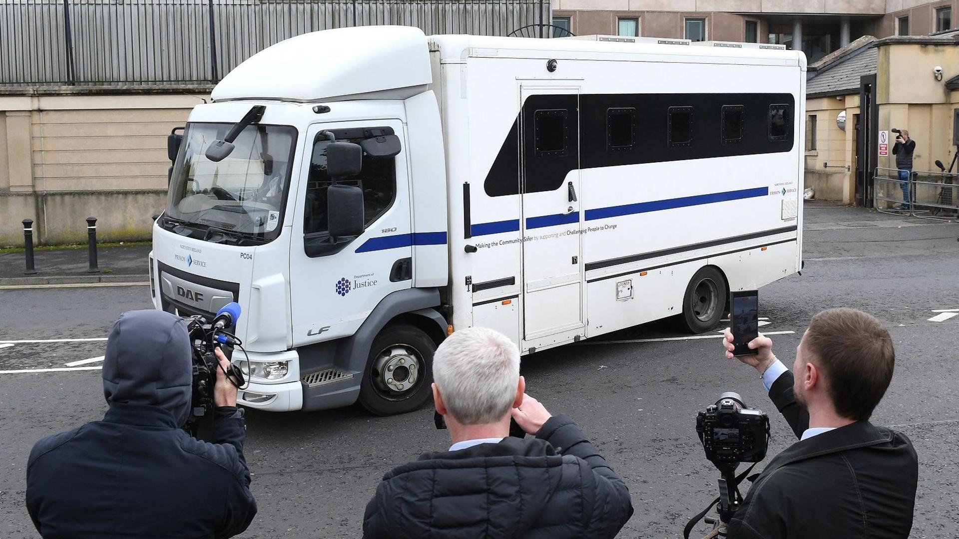 A white prison van on a road. The sky is overcast. Behind the van is a building. In the foreground, looking at the van, are three people with cameras.