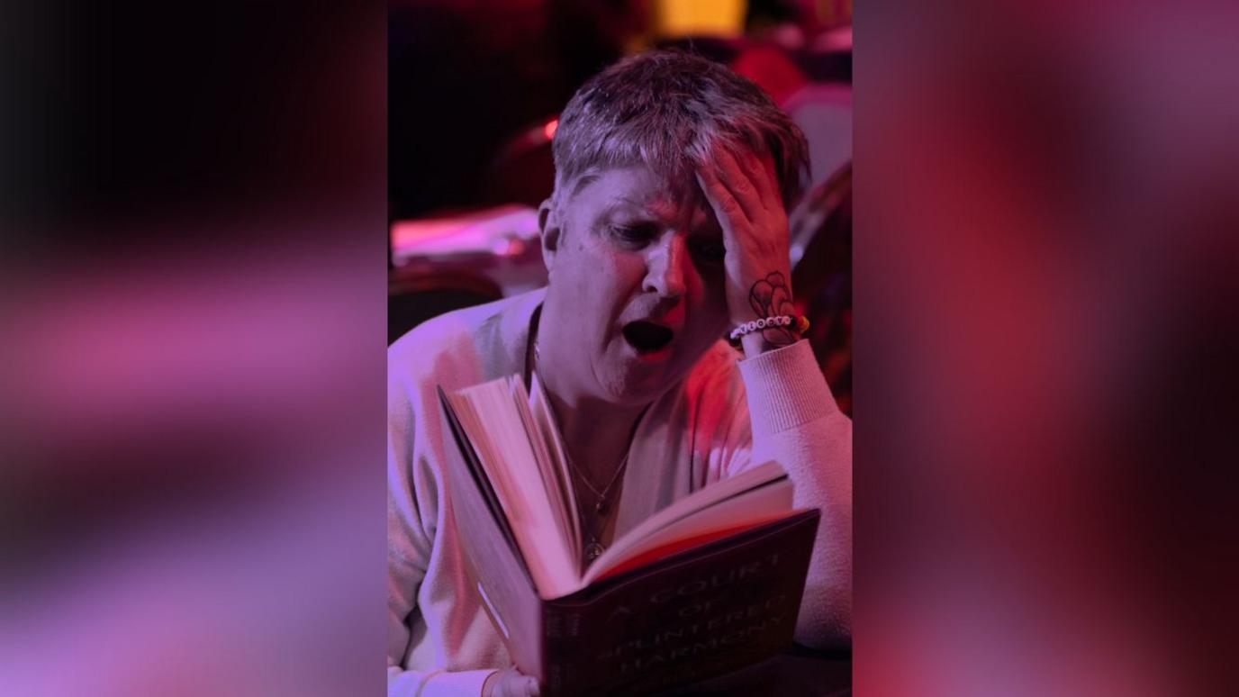 A woman with light, short hair is reading a book. She looks visibly shocked at what she is reading