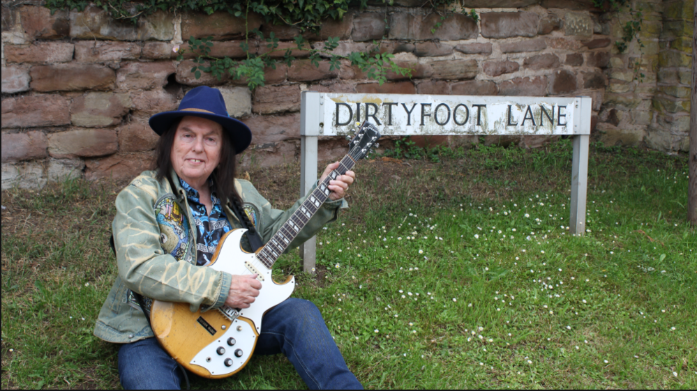A man is sitting on grass holding an electric guitar beside a street sign that reads “DIRTYFOOT LANE.” Behind them is an old stone wall with greenery growing along the top. The person is wearing a blue hat, a patterned shirt and a light-coloured jacket.
