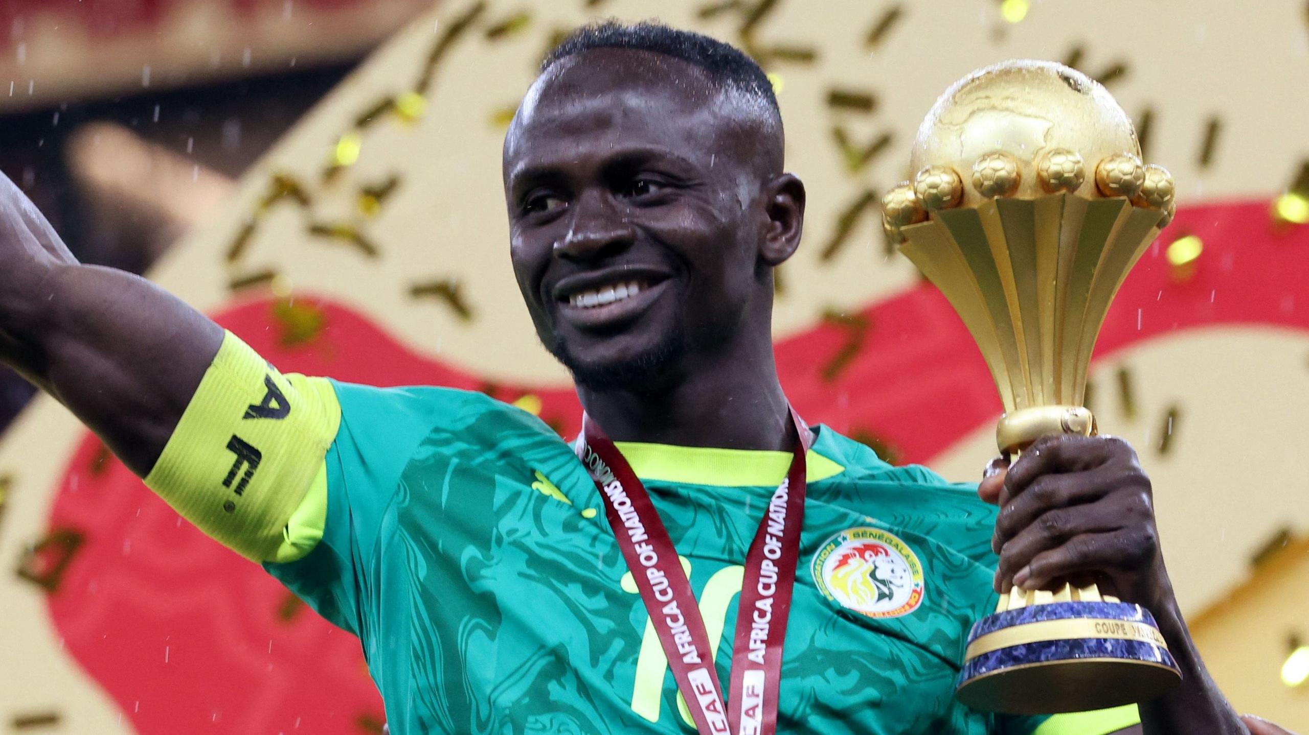 Sadio Mane, visible from chest up wearing a green Senegal shirt and with a yellow captain's armband on his right bicep, smiles as he holds the Africa Cup of Nations trophy in his left hand after the trophy presentation at Afcon 2025 in Morocco. He has a red ribbon around his neck, although the medal is out of shot, and golden ticker tape can be seen falling behind him