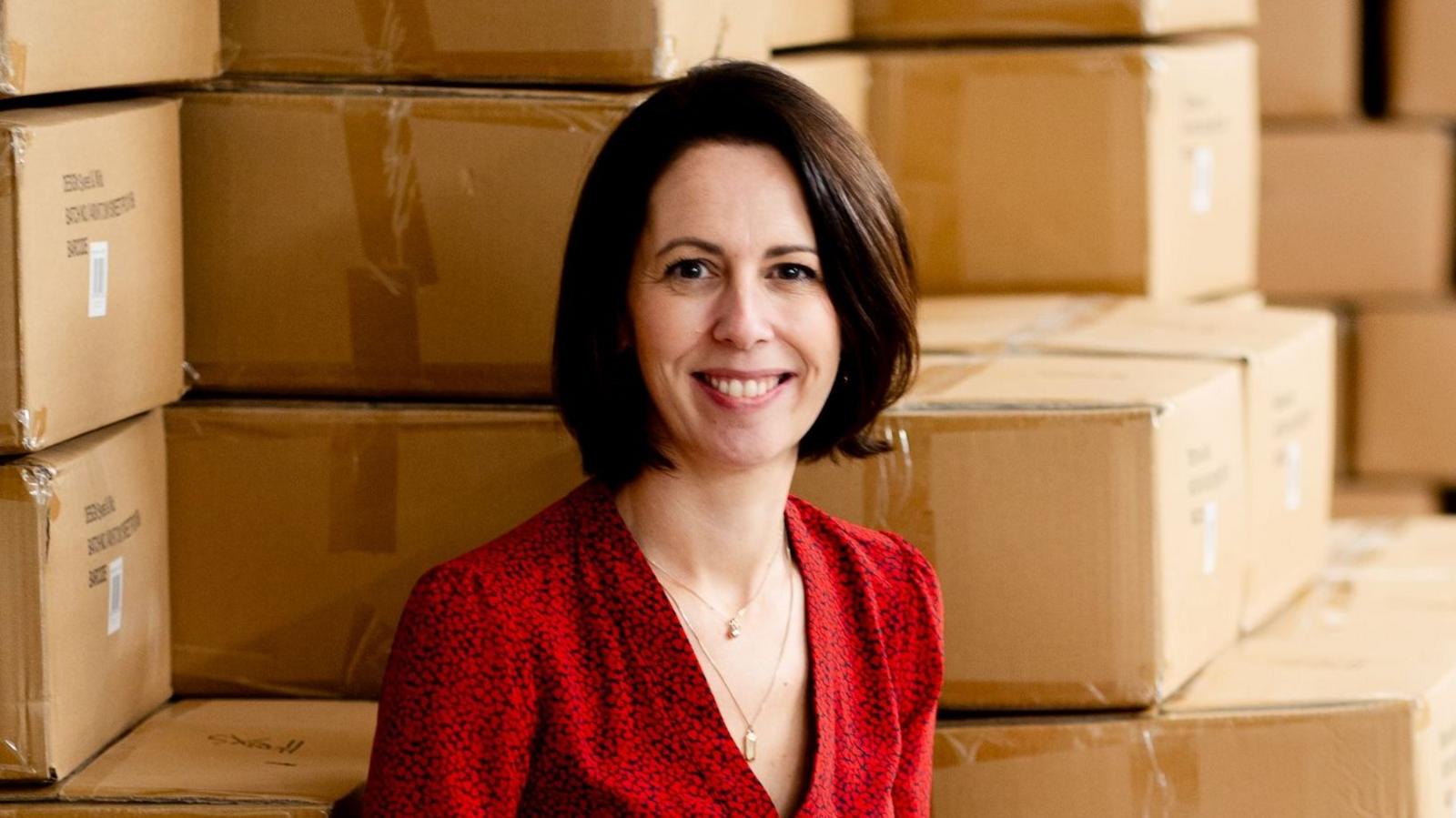 Irene Breen is pictured smiling. She has a dark brown bob haircut and is wearing a red shirt. She's sitting against stacks of boxes in a warehouse