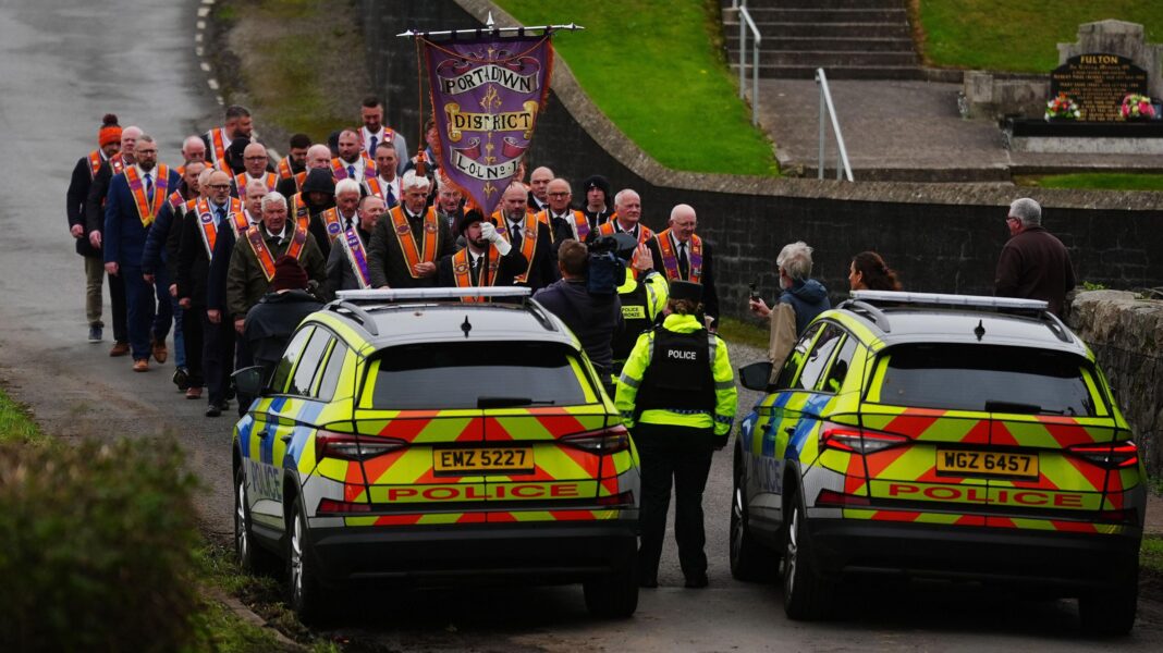 Two police cars are parked on a rural road and officers are standing beside them facing a small crowd of men wearing orange sashes. The men have a banner that says Portadown District Lol No 1. A cemetery is in the background.