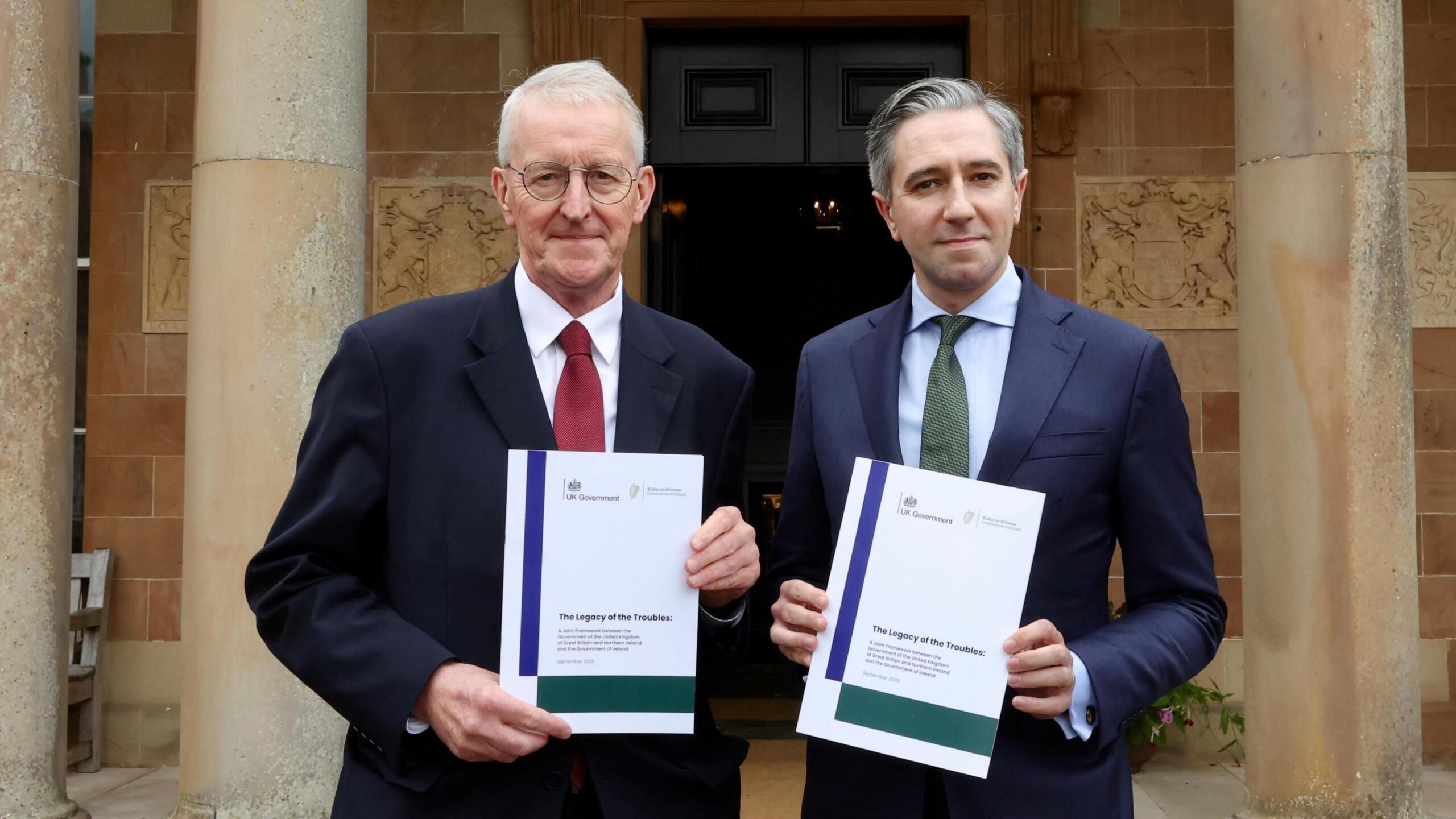 Hilary Benn, grey hair, spectacles, red tie, white shirt and Simon Harris, grey hair, blue jacket, blue shirt, green tie, hold up documents which are white, blue and green.